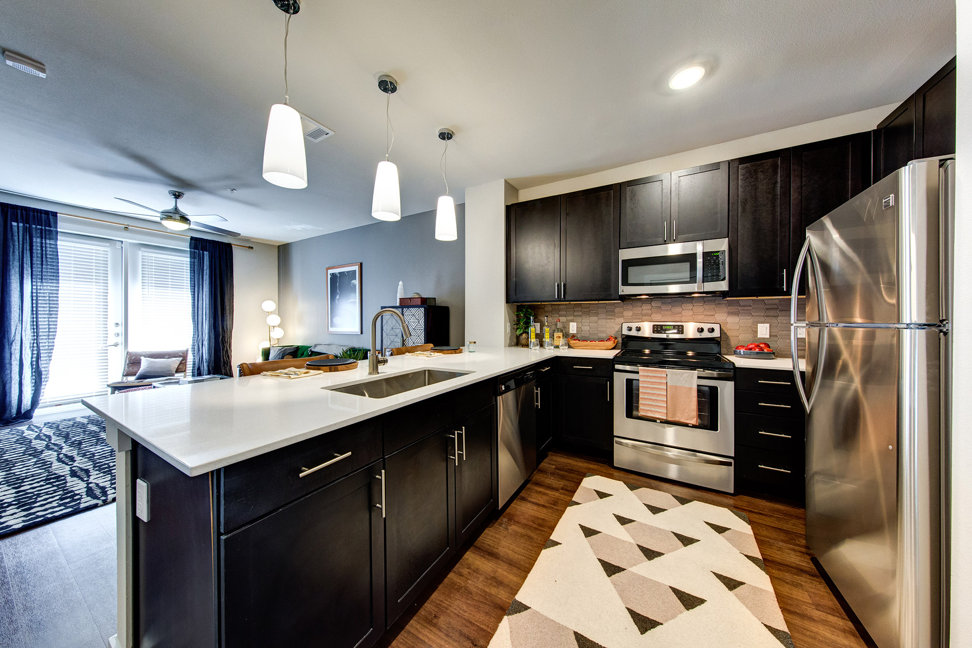 Kitchen with black cabinetry, wood-style floors, stainless steel appliances and quartz countertops that is open to the living room with double patio doors.